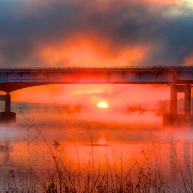 A vibrant sunrise glows behind a bridge spanning over a calm river, with mist rising from the water and silhouetted grass in the foreground. The sky is filled with dramatic orange and red hues.
