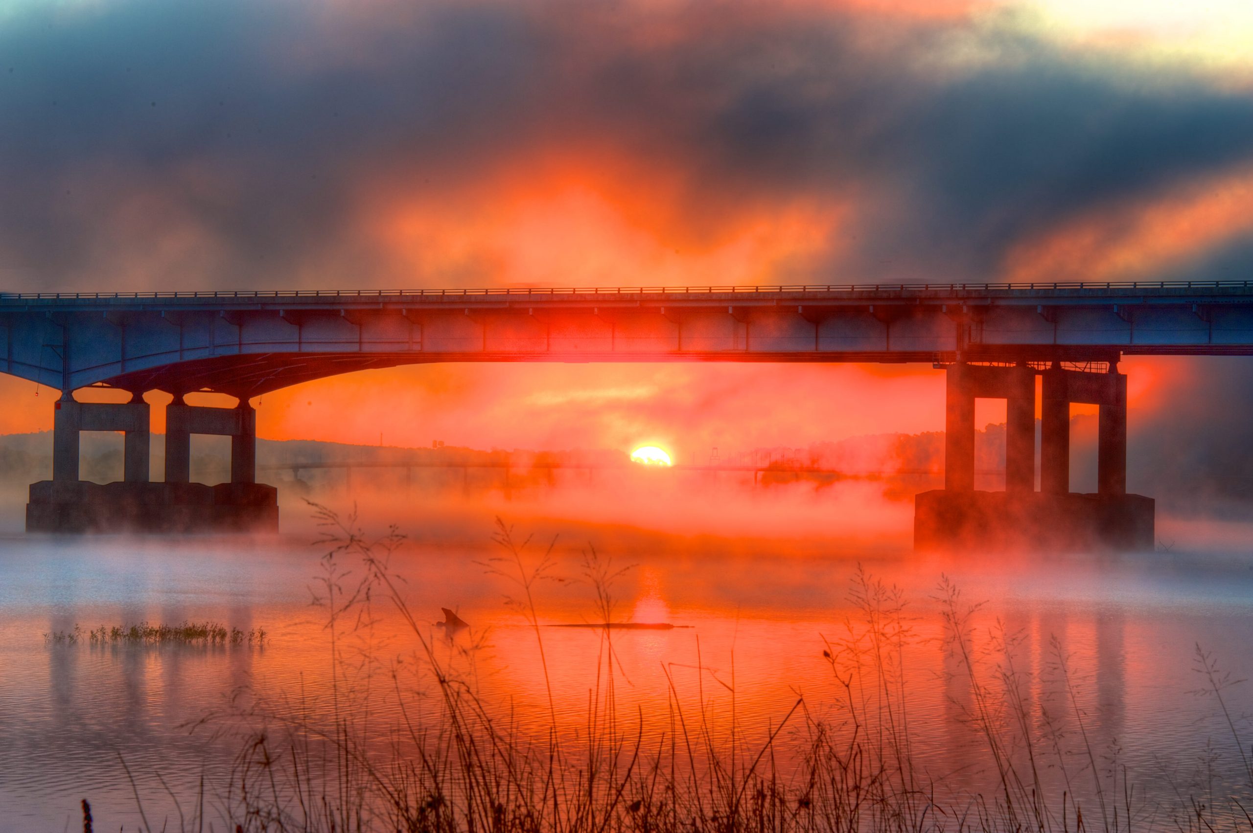 A vibrant sunrise glows behind a bridge spanning over a calm river, with mist rising from the water and silhouetted grass in the foreground. The sky is filled with dramatic orange and red hues.