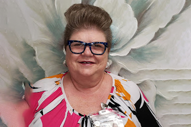 A smiling woman with short, styled hair and blue glasses wears a brightly patterned top and a name badge, standing in front of a large, floral mural.