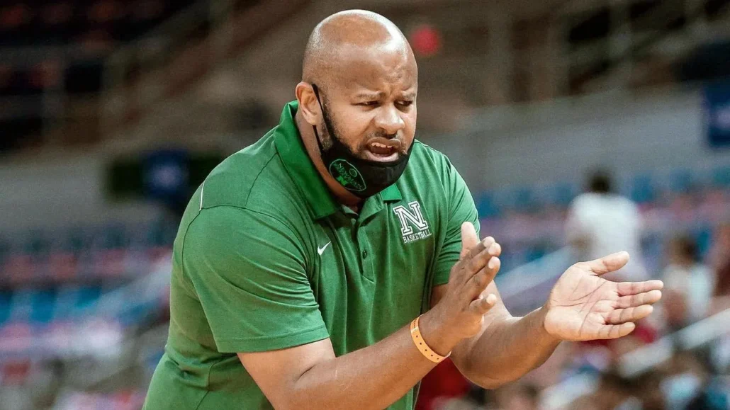 A basketball coach in a green polo shirt claps his hands and appears to be giving instructions, with a mask pulled down under his chin in an indoor sports arena.
