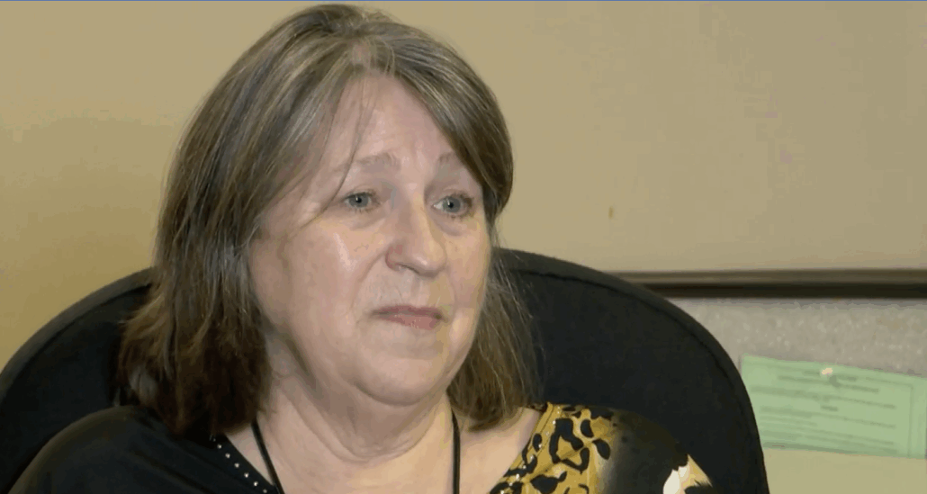 A woman with shoulder-length brown and gray hair sits in an office chair, wearing a black and yellow patterned top. She has a neutral expression and appears to be listening or talking. A beige wall and papers are visible in the background.