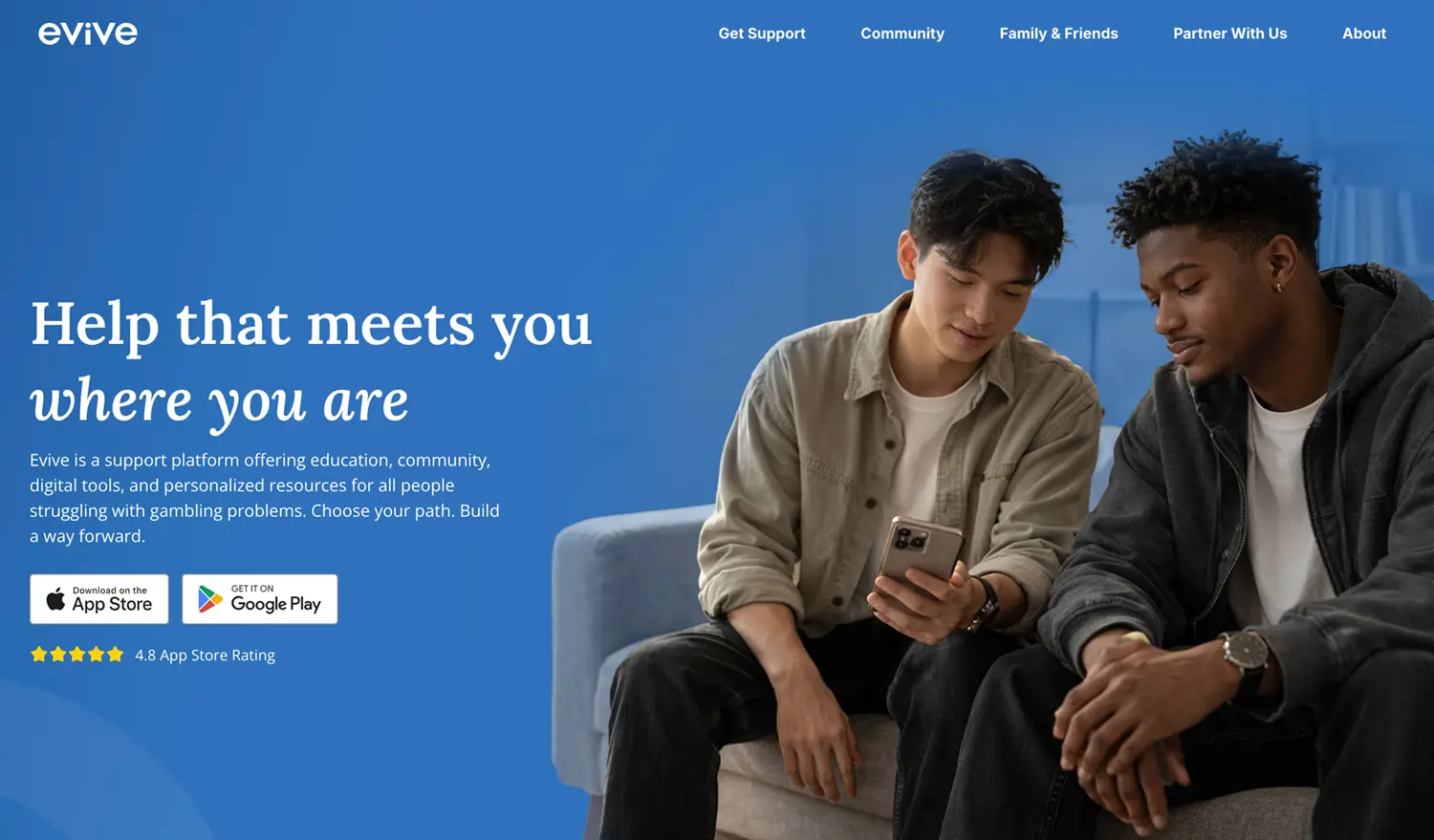 Two young men sit on a couch looking at a smartphone together, smiling. The background is blue with white text promoting Evive, a support platform for people struggling with gambling. App store download buttons are visible.